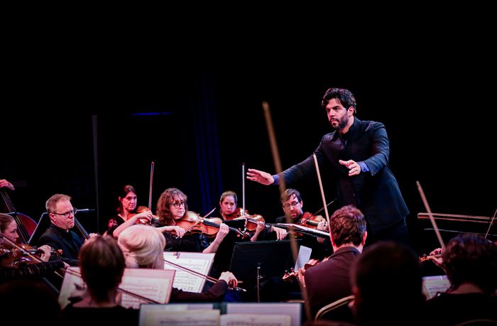 Domingo Hindoyan stood with his arms lifted towards members of the strings section in the orchestra who are wearing black and playing their instruments. There is cream hue lighting.