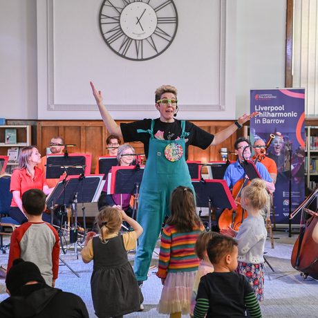 Present Claire Henry standing in the centre wearing green dungarees with their arms in the air talking to a group of children who are all standing with their backs to the camera, members of the Orchestra are sitting behind Claire wearing bright coloured shirts