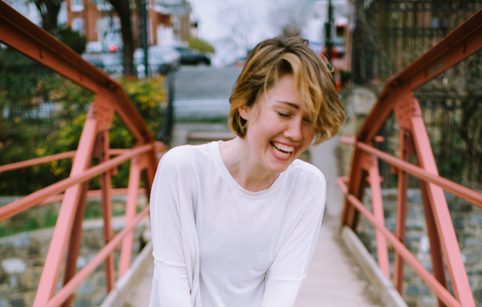 Caroline Shaw with short light brown hair, wearing a white shirt and smiling with their eyes looking down