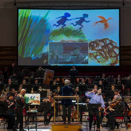 Members of the orchestra performing on stage with a live artist painting a forest scene on a screen