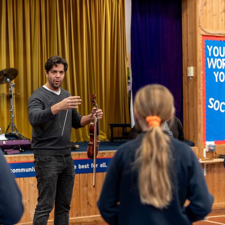 Royal Liverpool Philharmonic Orchestra Chief Conductor Domingo Hindoyan holds a violin while talking to a group of schoolchildren wearing blue uniform in a school hall