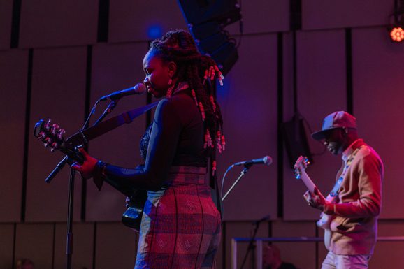 A young female musician with black braided hair and patterned trousers plays the guitar on stage in the Music Room with another musician behind her