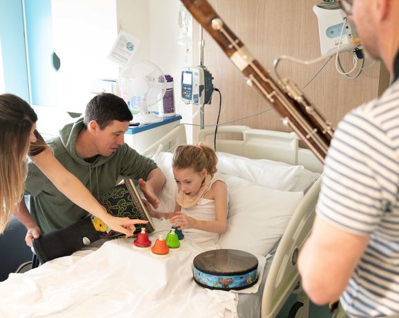 Young girl in a hospital bed playing with instruments with her parents and a Liverpool Philharmonic musician next to her