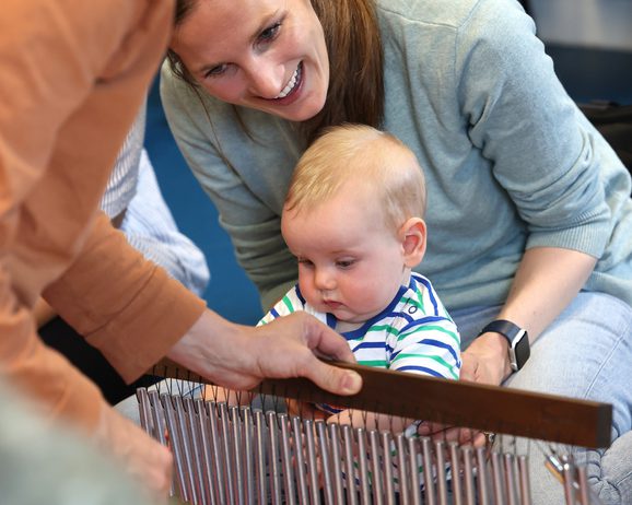 A young baby sat on the floor with their smiling mother playing with a percussion instrument