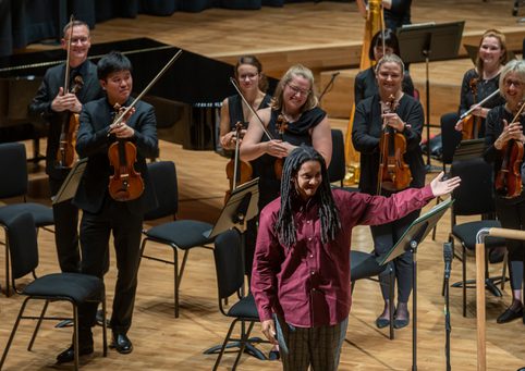 Musicians dressed all in black on their feet, Rushworth Composition Prize winner Nneka Cummins wearing a red shirt stands at the front of the stage with her arm stretched out