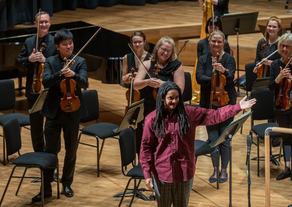 Musicians dressed all in black on their feet, Rushworth Composition Prize winner Nneka Cummins wearing a red shirt stands at the front of the stage with her arm stretched out