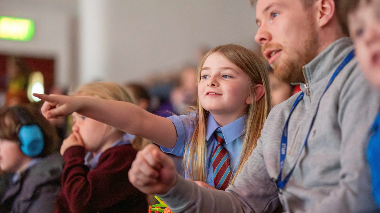 Girl with blonde hair in school uniform sat in Liverpool Philharmonic Hall pointing at the stage and asking her teacher a question