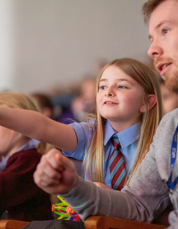 Girl with blonde hair in school uniform sat in Liverpool Philharmonic Hall pointing at the stage and asking her teacher a question