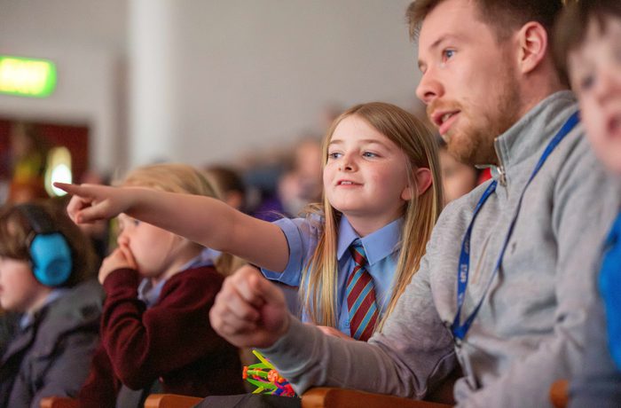 Girl with blonde hair in school uniform sat in Liverpool Philharmonic Hall pointing at the stage and asking her teacher a question