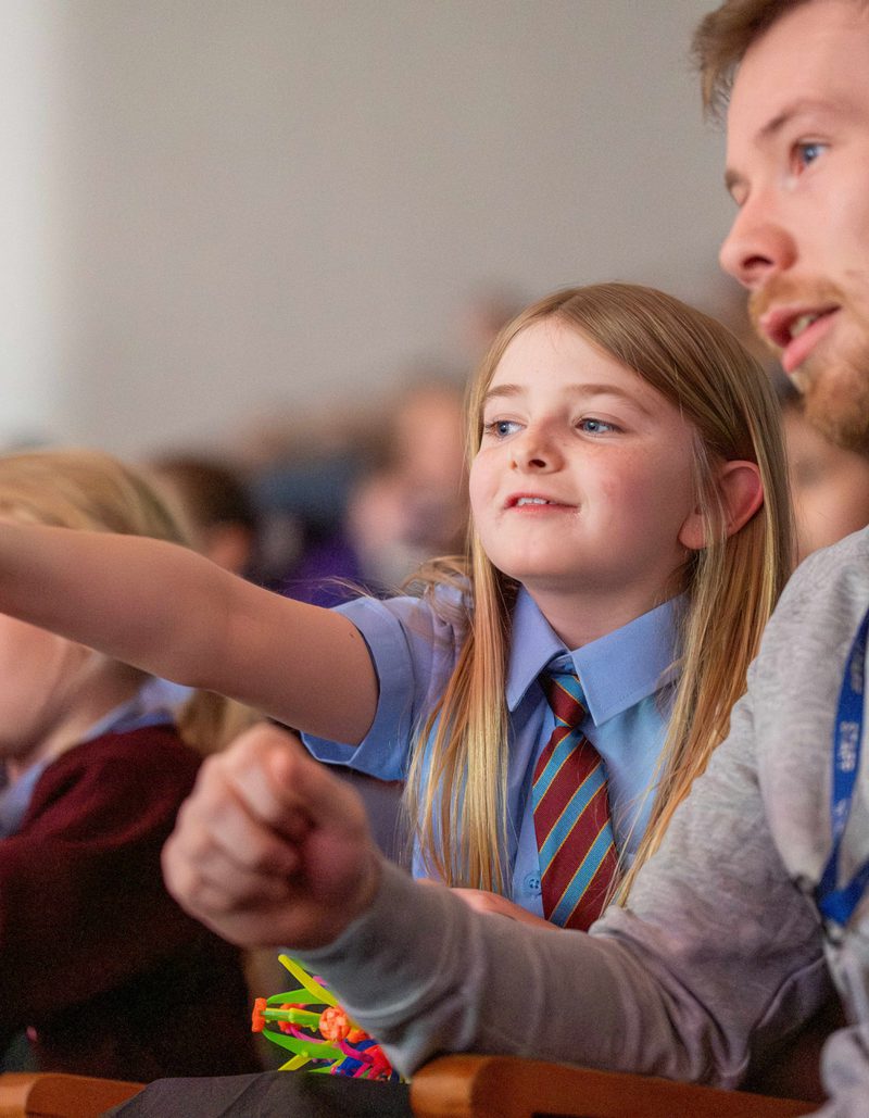 Girl with blonde hair in school uniform sat in Liverpool Philharmonic Hall pointing at the stage and asking her teacher a question