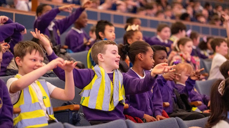A group of schoolchildren in purple uniforms and high-vis jackets clapping and laughing in Liverpool Philharmonic Hall