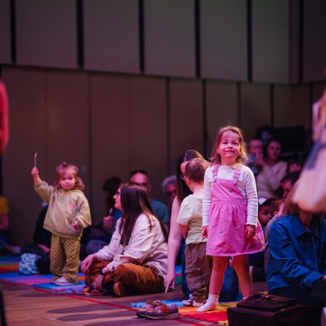 A crowd of parents and toddlers sat on the floor in the Music Room with one little girl standing up, smiling and staring at the performers