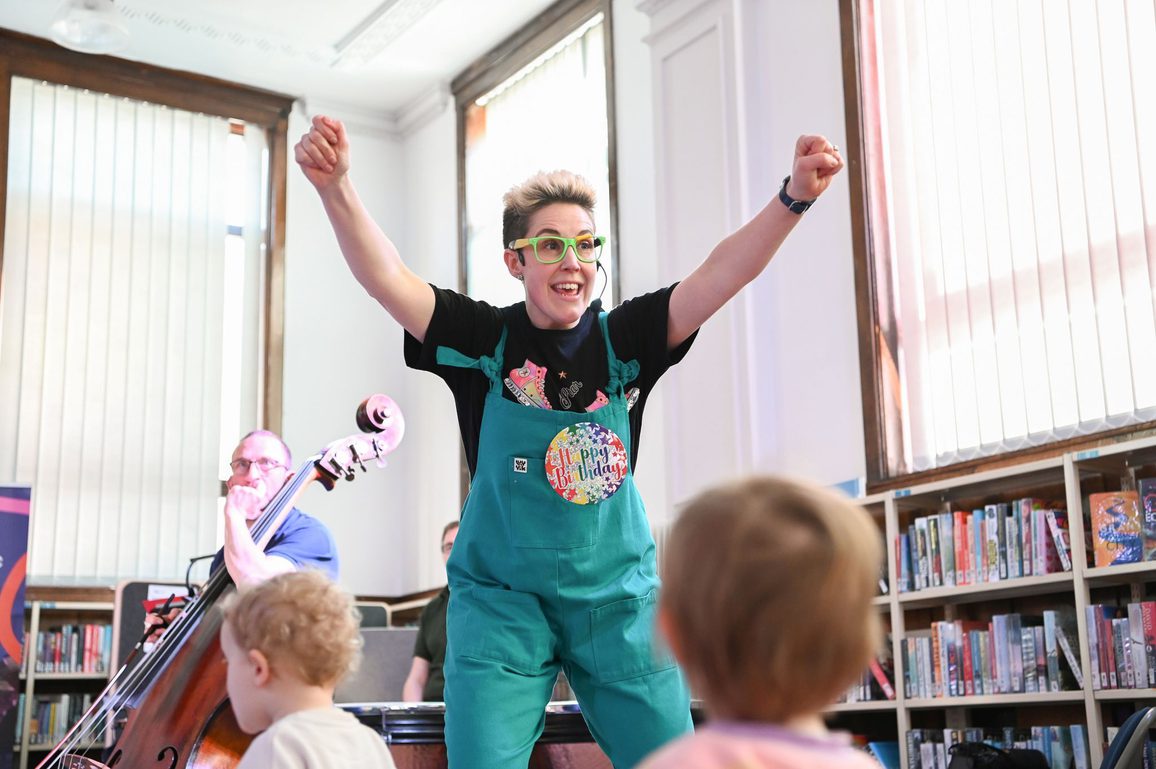 Under 5s concert presenter Claire Henry standing with her arms in the air in front of the musicians with toddlers in front of her looking at the performers