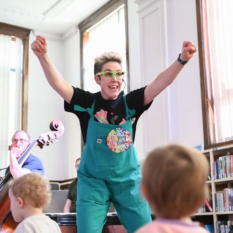 Under 5s concert presenter Claire Henry standing with her arms in the air in front of the musicians with toddlers in front of her looking at the performers
