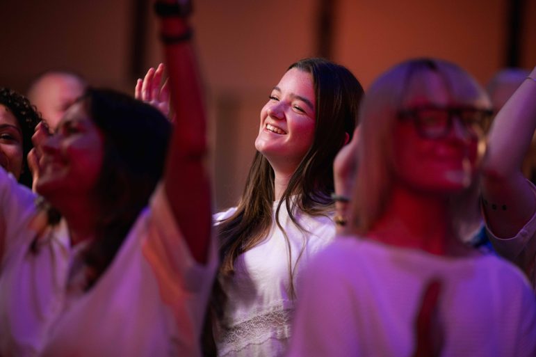 Young woman in a white top standing and smiling at the stage with other audience members in the foreground with their hands in the air