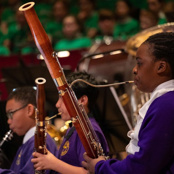 A young girl in a purple school uniform on stage playing the bassoon, with two boys in the background also in purple school uniform and playing woodwind instruments