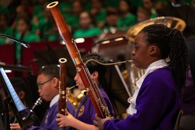 A young girl in a purple school uniform on stage playing the bassoon, with two boys in the background also in purple school uniform and playing woodwind instruments