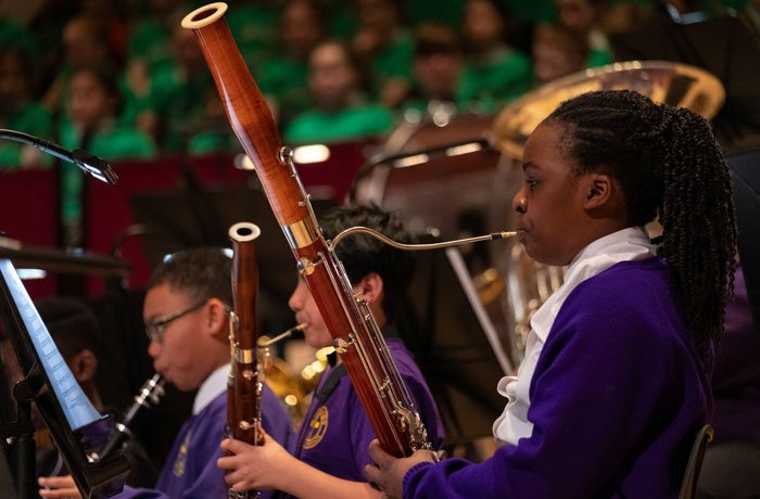 A young girl in a purple school uniform on stage playing the bassoon, with two boys in the background also in purple school uniform and playing woodwind instruments