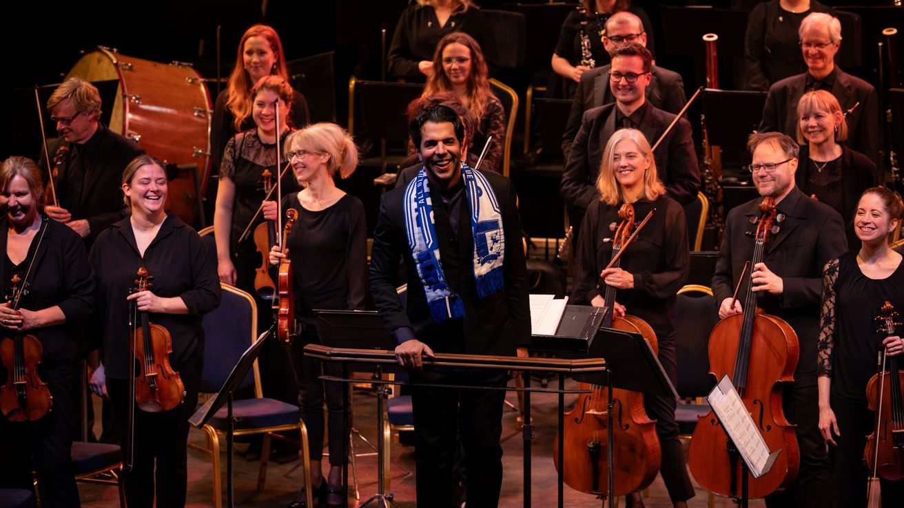 Chief Conductor Domingo Hindoyan standing in front of the Orchestra wearing a Barrow football scarf and smiling at the audience