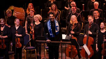 Chief Conductor Domingo Hindoyan standing in front of the Orchestra wearing a Barrow football scarf and smiling at the audience