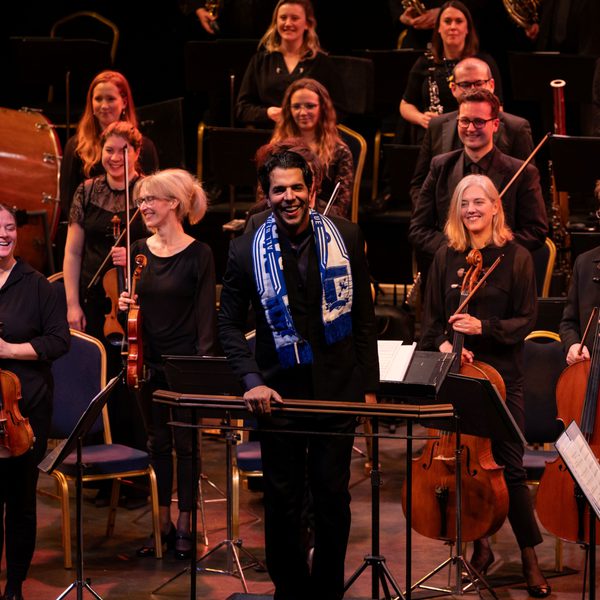 Chief Conductor Domingo Hindoyan standing in front of the Orchestra wearing a Barrow football scarf and smiling at the audience