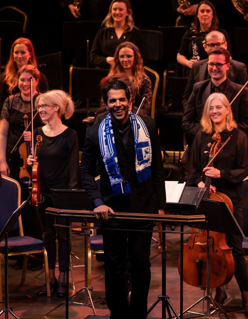 Chief Conductor Domingo Hindoyan standing in front of the Orchestra wearing a Barrow football scarf and smiling at the audience