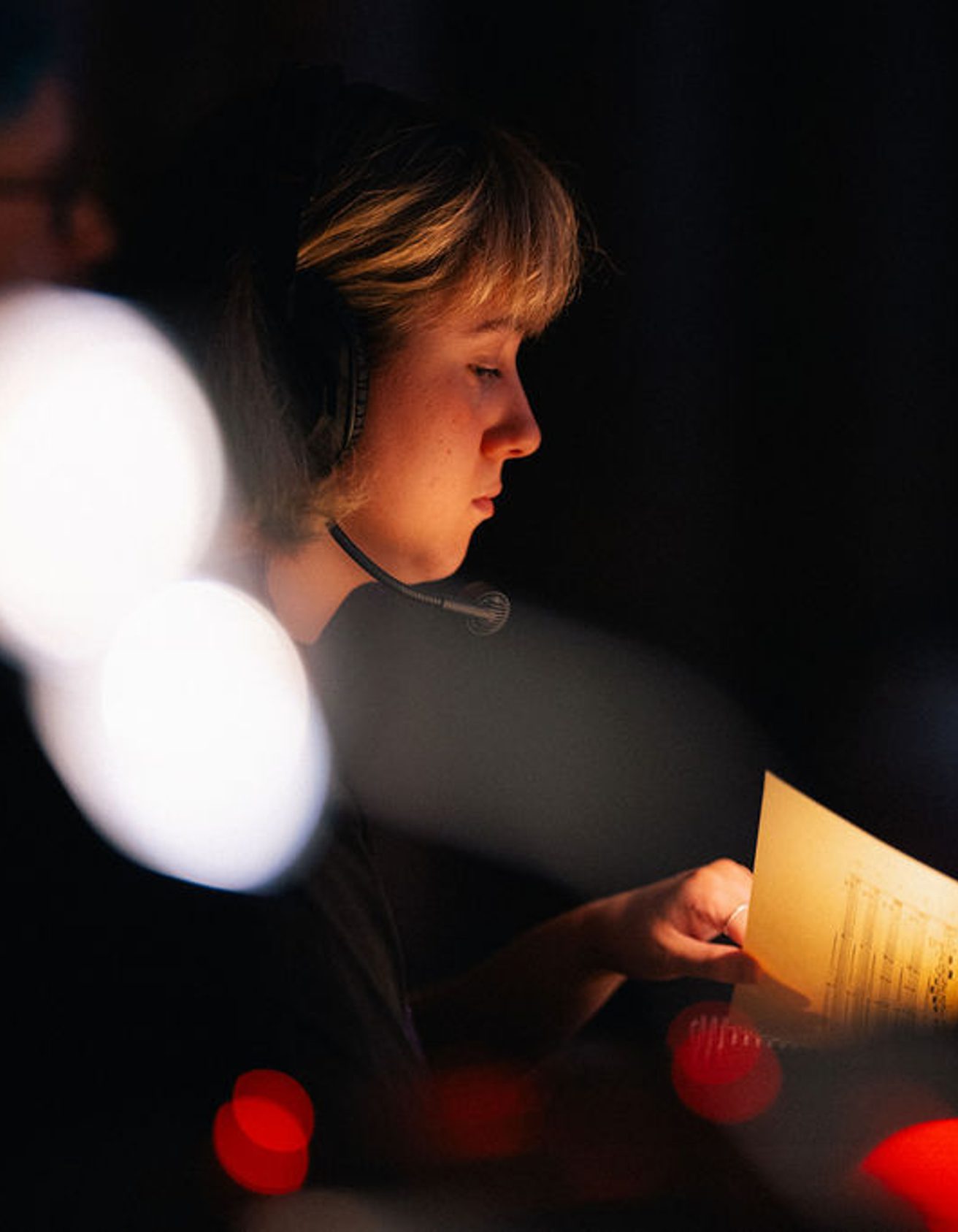 Girl with short blonde hair wearing a headset sits backstage at a performance looking at a musical score