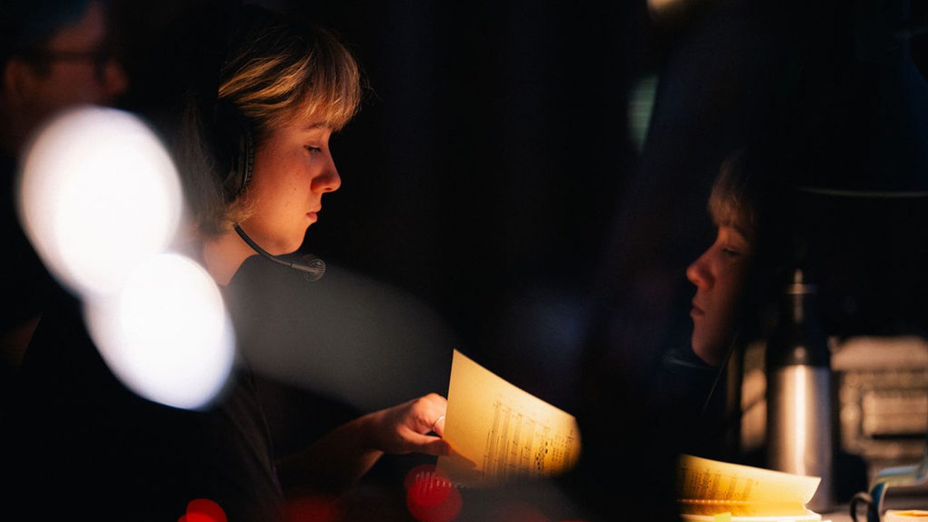 Girl with short blonde hair wearing a headset sits backstage at a performance looking at a musical score