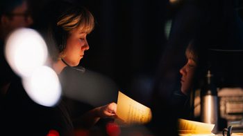 Girl with short blonde hair wearing a headset sits backstage at a performance looking at a musical score
