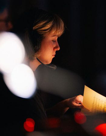 Girl with short blonde hair wearing a headset sits backstage at a performance looking at a musical score