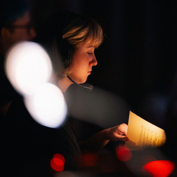 Girl with short blonde hair wearing a headset sits backstage at a performance looking at a musical score