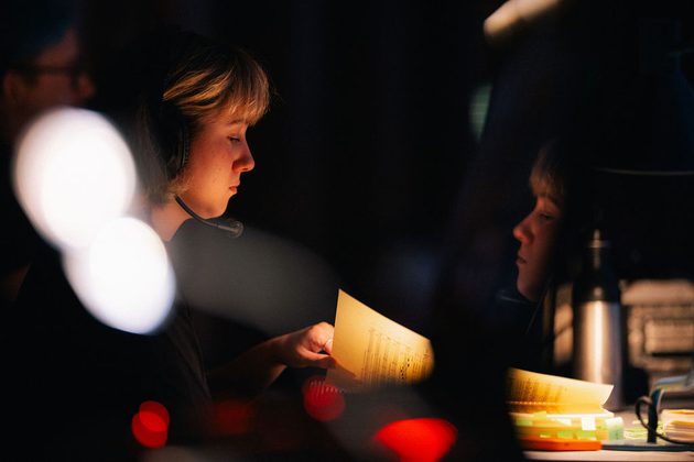 Girl with short blonde hair wearing a headset sits backstage at a performance looking at a musical score