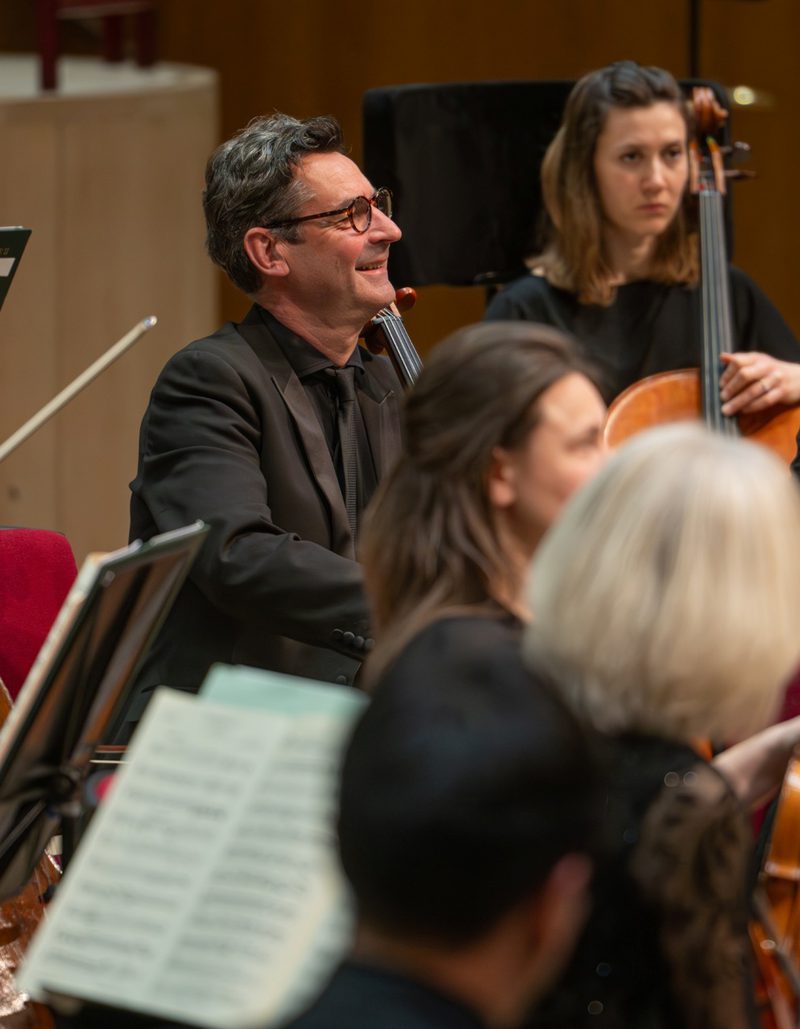 Cello section of the orchestra wearing all black and smiling playing their instruments