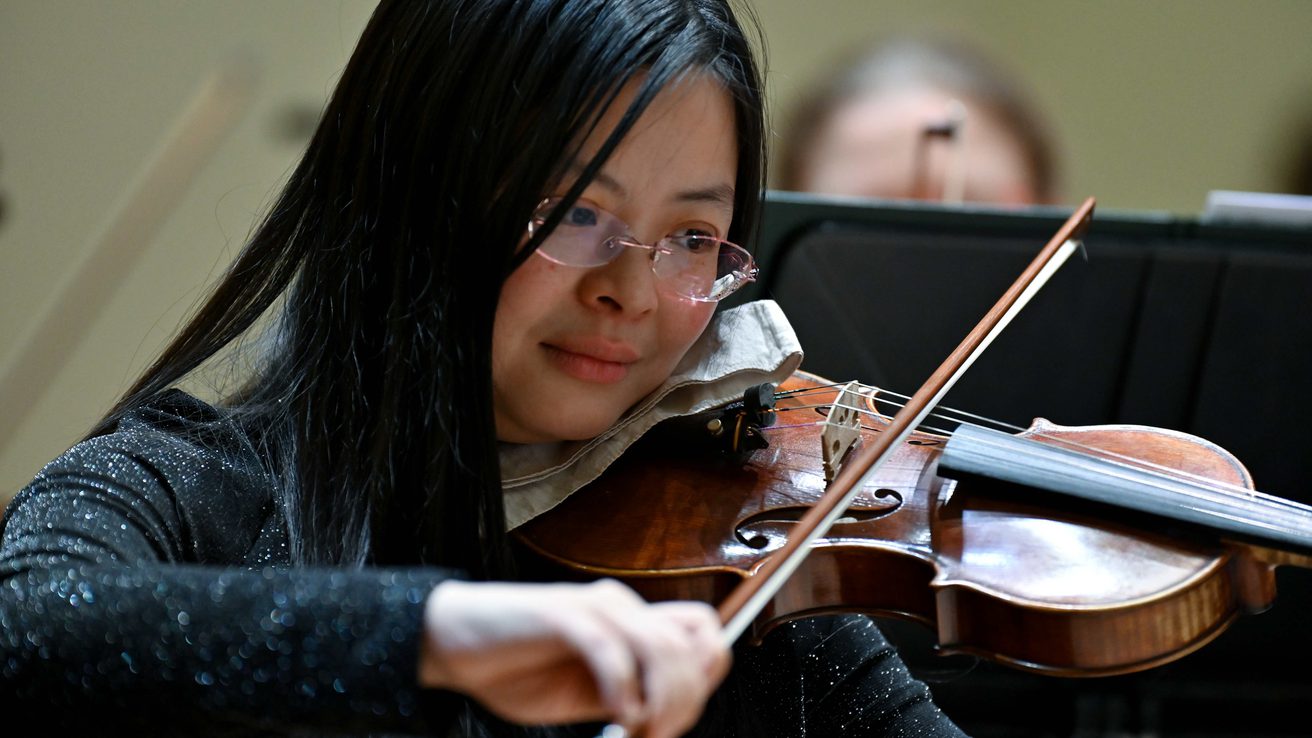A member of the Orchestra with glasses and short dark playing the violin