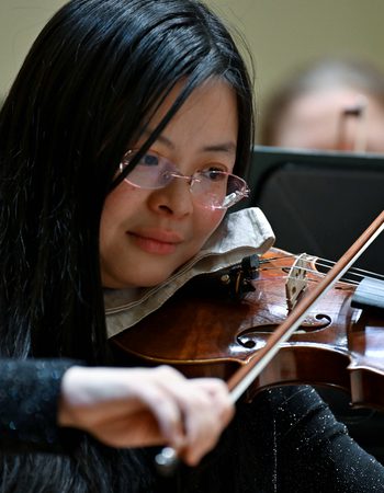 A member of the Orchestra with glasses and short dark playing the violin