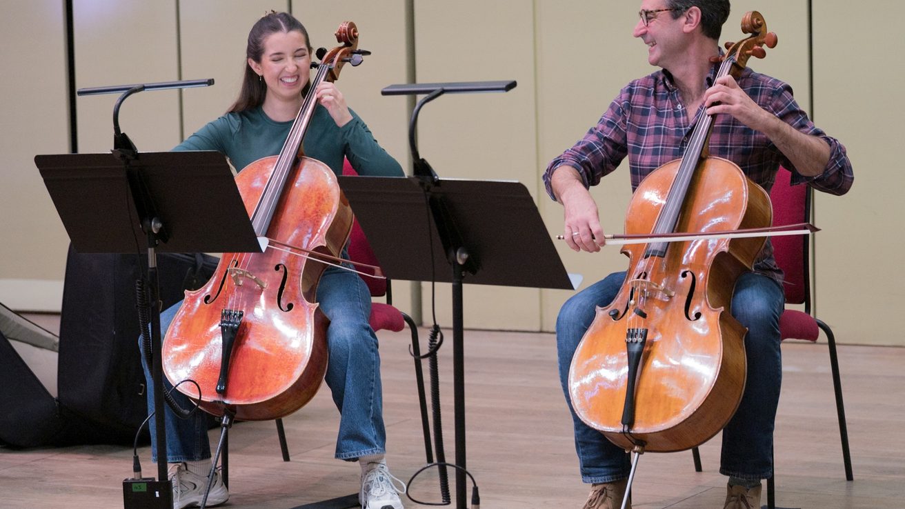 two musicians smiling while playing the cello with black music stands in front of them