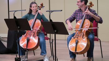 two musicians smiling while playing the cello with black music stands in front of them