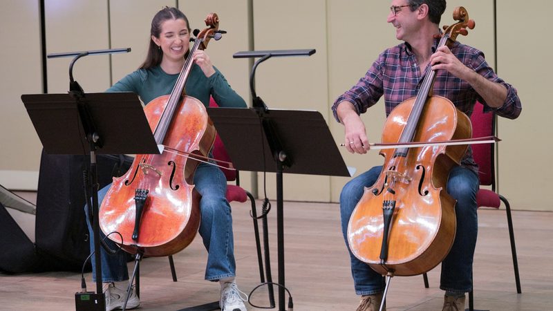 two musicians smiling while playing the cello with black music stands in front of them