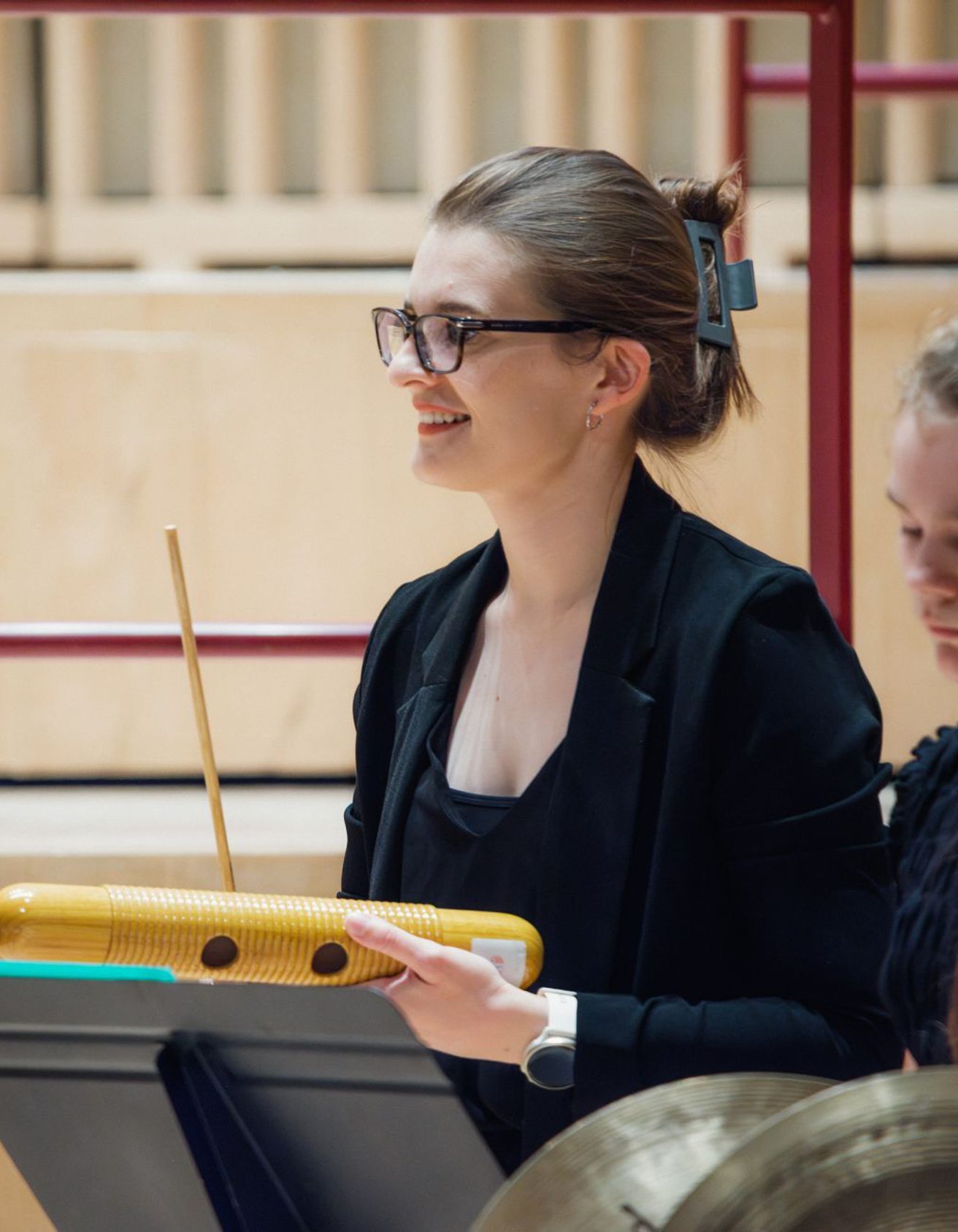 Young girl wearing black clothing and black glasses holding a wooden guiro scraper and smiling to the left