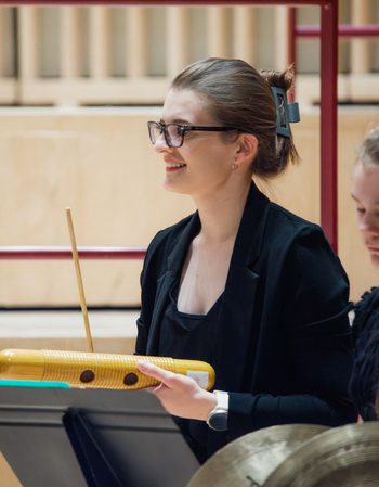 Young girl wearing black clothing and black glasses holding a wooden guiro scraper and smiling to the left