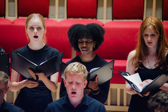 three girls stood in a row wearing black, looking down at an opened black book whilst singing