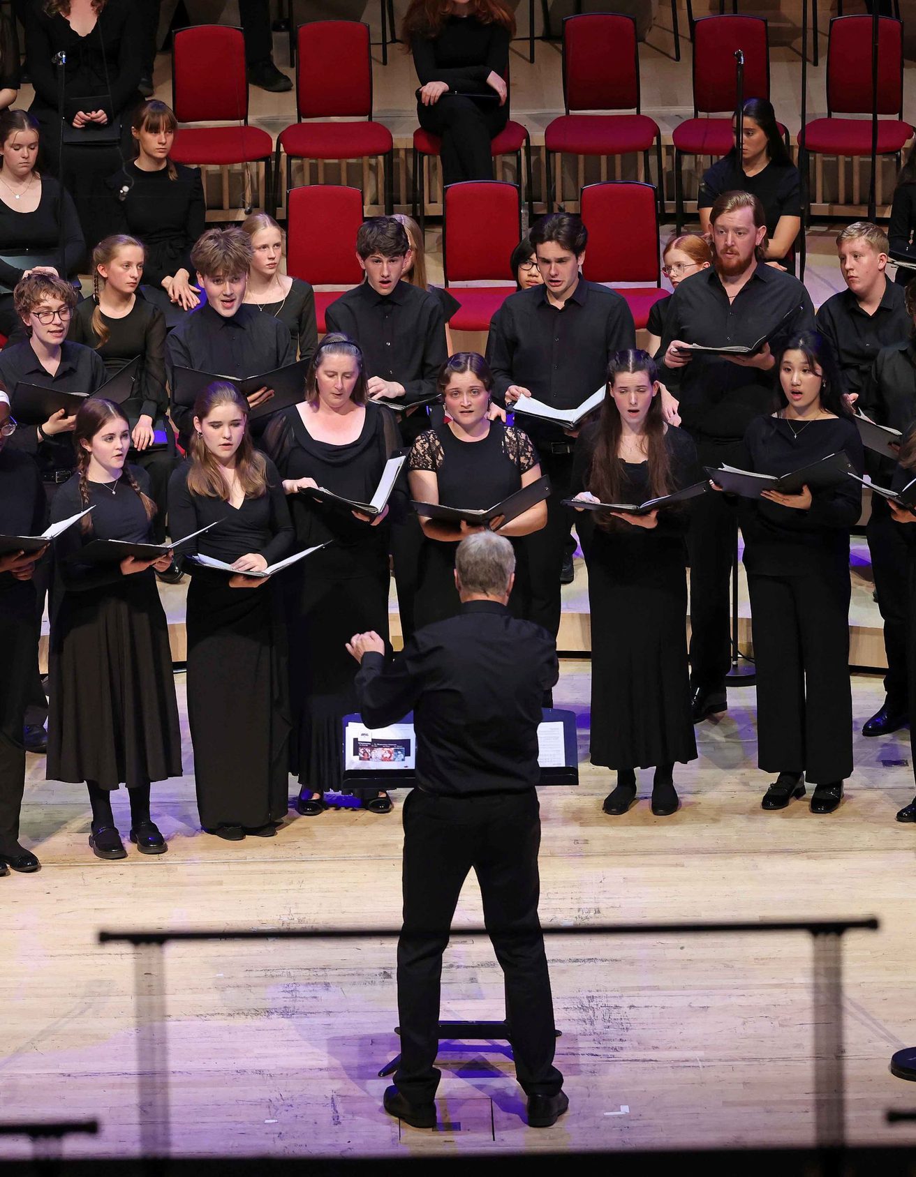 All members of the Youth Choir on stage wearing black and holding an opened black book, they're looking at the conductor who's at the centre of the stage wearing black with their left arm slightly raised