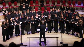 All members of the Youth Choir on stage wearing black and holding an opened black book, they're looking at the conductor who's at the centre of the stage wearing black with their left arm slightly raised