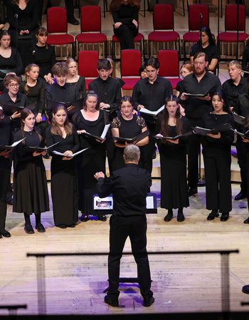All members of the Youth Choir on stage wearing black and holding an opened black book, they're looking at the conductor who's at the centre of the stage wearing black with their left arm slightly raised