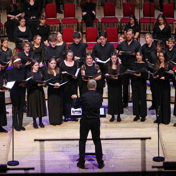 All members of the Youth Choir on stage wearing black and holding an opened black book, they're looking at the conductor who's at the centre of the stage wearing black with their left arm slightly raised