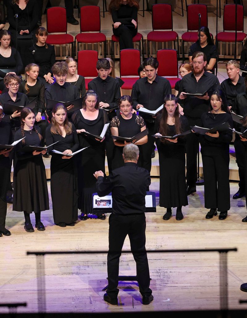 All members of the Youth Choir on stage wearing black and holding an opened black book, they're looking at the conductor who's at the centre of the stage wearing black with their left arm slightly raised