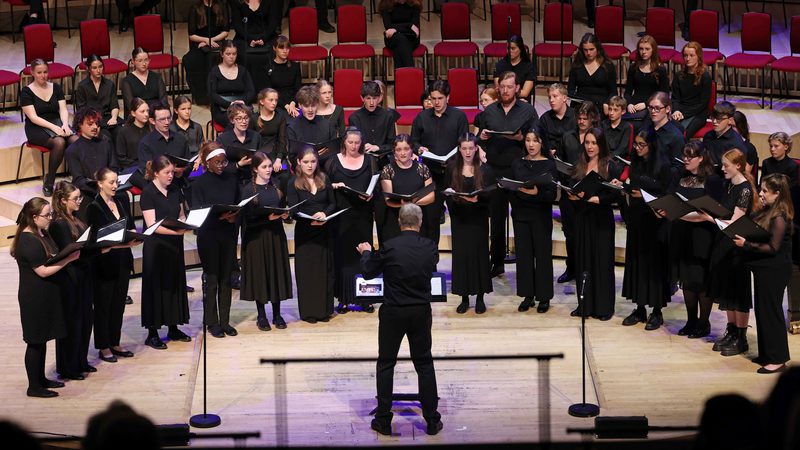 All members of the Youth Choir on stage wearing black and holding an opened black book, they're looking at the conductor who's at the centre of the stage wearing black with their left arm slightly raised