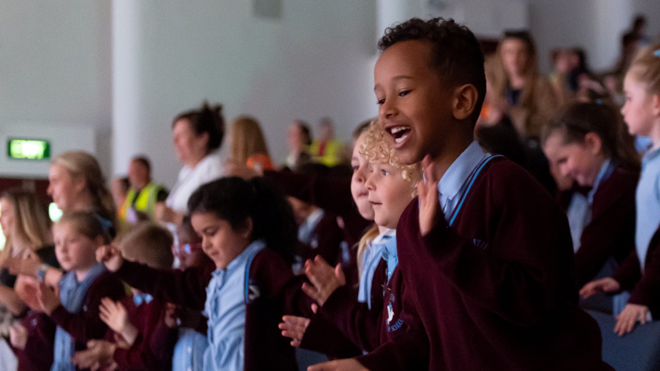 Young audience members wearing blue and burgundy school uniform smiling and clapping