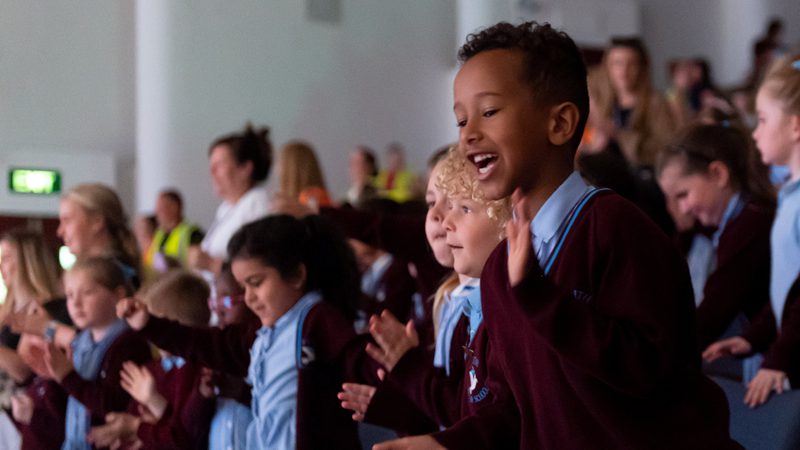 Young audience members wearing blue and burgundy school uniform smiling and clapping