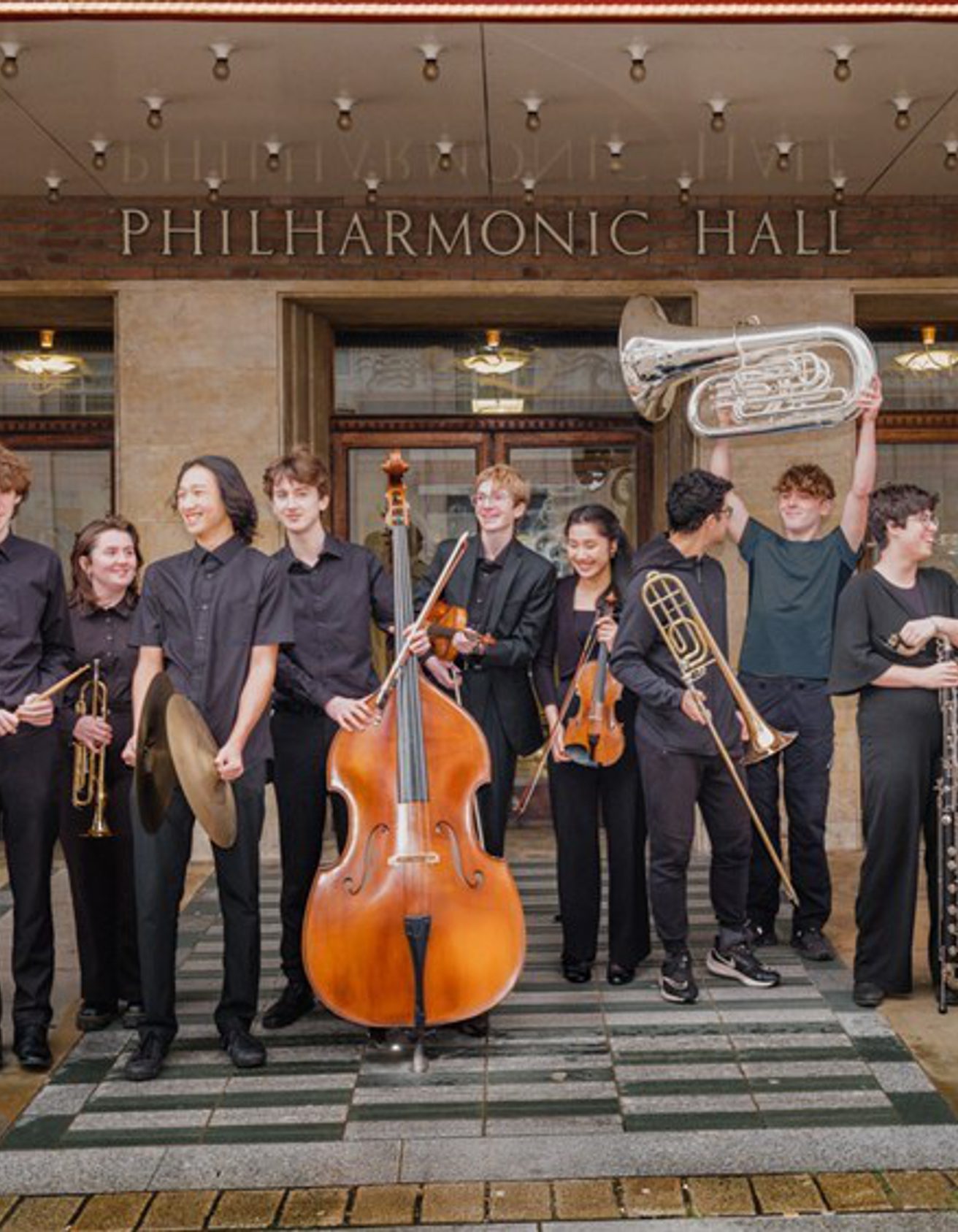Members of the Youth Company standing in a row outside Liverpool Philharmonic Hall wearing black and holding their instruments